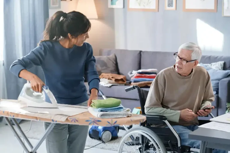 volunteer-ironing-clothes-talking-senior-man-who-using-wheelchair-she-helping-him-with-chores-room
